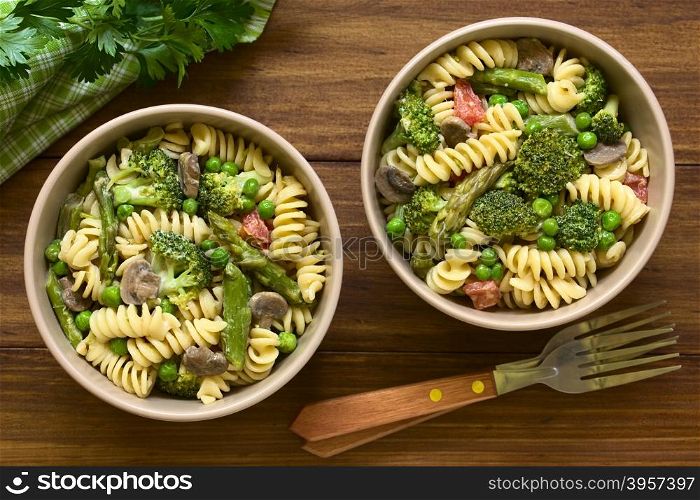 Pasta primavera with green asparagus, pea, broccoli, mushroom and tomato in cream sauce served in bowls, photographed overhead on dark wood with natural light