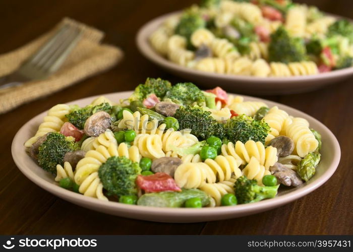 Pasta primavera with green asparagus, pea, broccoli, mushroom and tomato in cream sauce served on plates, photographed on dark wood with natural light (Selective Focus, Focus in the middle of the pasta on the first plate)