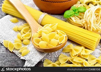 Pasta seashells in spoon, pipe rigate in a bowl, spaghetti, tagliatelle, farfalle, trenette on burlap on a wooden board background