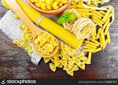 Pasta seashells in spoon, pipe rigate in a bowl, spaghetti, tagliatelle, penne, farfalle, trenette on napkin of burlap on wooden board background from above