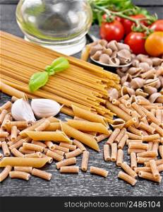 Pasta whole grain and rye pipe rigate, spaghetti, penne; seashells, rigatoni, tomatoes on a branch, garlic, vegetable oil in a decanter on black wooden board background