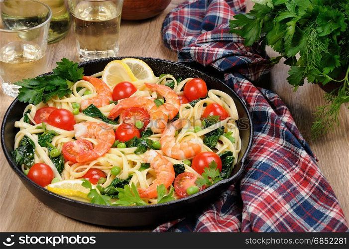Pasta with fried prawns, peas, tomatoes and spinach in a frying pan, on a table with cider glasses.