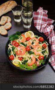 Pasta with fried prawns, peas, tomatoes and spinach in a frying pan, on a table with cider glasses.