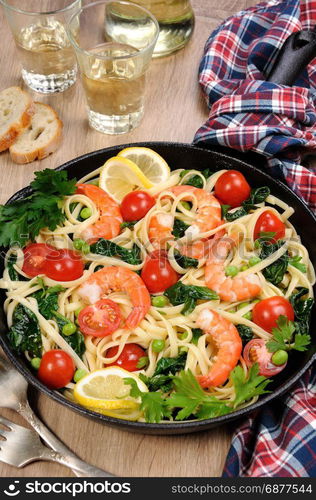 Pasta with fried prawns, peas, tomatoes and spinach in a frying pan, on a table with cider glasses.