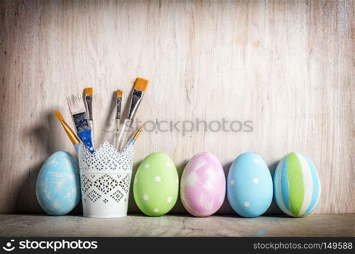 Pastel Easter eggs and brushes in a rustic cup on wooden desk.. Pastel Easter eggs and brushes in a rustic cup