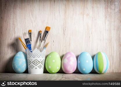 Pastel Easter eggs and brushes in a rustic cup on wooden desk.. Pastel Easter eggs and brushes in a rustic cup