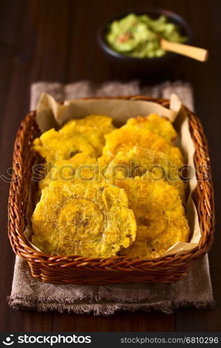 Patacon or toston, fried and flattened pieces of green plantains, a traditional snack or accompaniment in the Caribbean, guacamole in the back, photographed on dark wood with natural light (Selective Focus, Focus on the first slice)