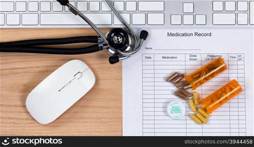 Patient medication record form with stethoscope, medication container, capsules, computer keyboard and mouse on wooden desktop. Mouse and keyboard are generic brand items for display purposes only.