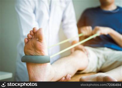 Patient use resistance band stretching out his leg with physical therapist helps in clinic room.