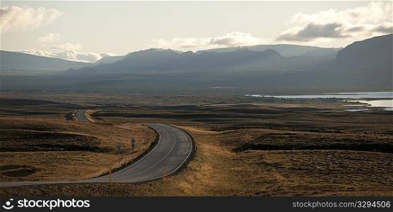 Paved highway meandering through dry farmland towards misty mountains