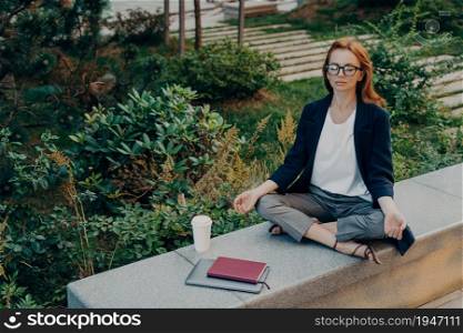 Peaceful calm redhead business woman doing yoga with closed eyes outdoor, relaxing in lotus pose during remote work in park, female relieving stress while working on laptop outside. Meditation concept. Peaceful calm redhead business woman doing yoga with closed eyes outdoor