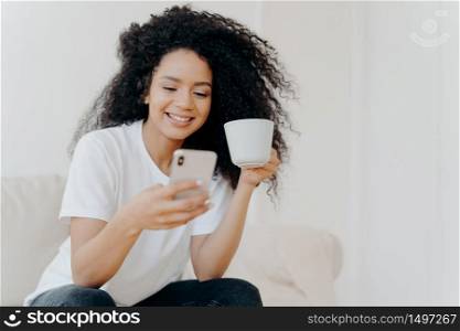 Peaceful curly woman scrolling social networks via modern mobile phone, watches funny videos, views photos in social networks, drinks coffee, poses on couch in living room, gets high paid job offer