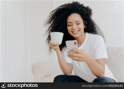 Peaceful curly woman scrolling social networks via modern mobile phone, watches funny videos, views photos in social networks, drinks coffee, poses on couch in living room, gets high paid job offer