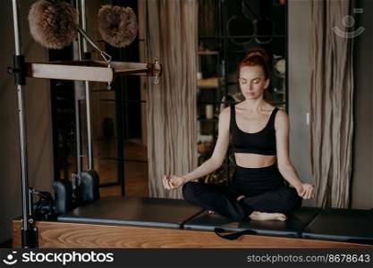 Peaceful female pilates instructor sitting in yoga lotus position on Cadillac reformer, trying to relax after intensive stretching exercises in pilates studio. Meditation and yoga concept. Young redhead woman in sportswear sitting in lotus position on trapeze table trying to relax