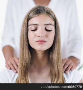 Peaceful girl sitting with her eyes closed and receiving Reiki healing treatment of female Reiki therapist. Reiki Practitioner standing and massages shoulders of beautiful teenage patient. Alternative therapy concept