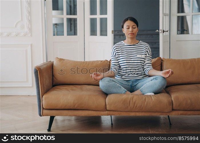 Peaceful hispanic woman is practicing yoga at home. Lady is sitting alone on sofa in lotus asana and meditating in living room. Stress relief and relaxation. Contemplation and mindfulness.. Lady sitting on sofa in lotus asana and meditating in living room. Stress relief and relaxation.