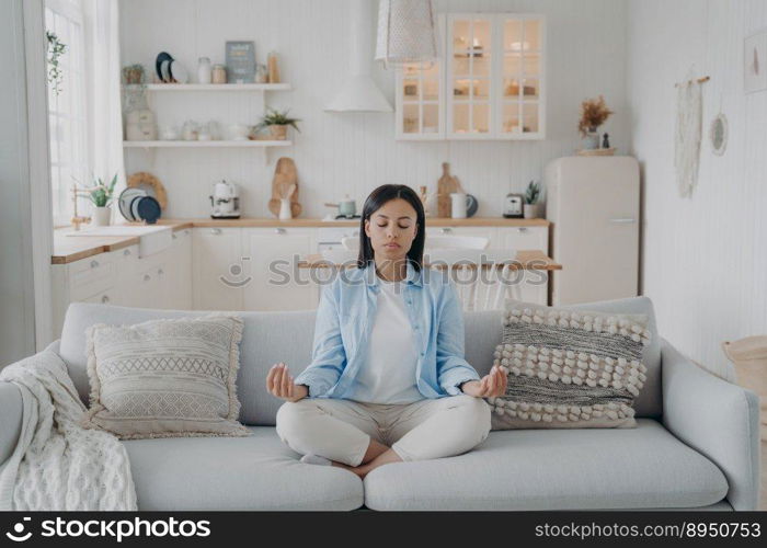 Peaceful mind, zen and concentration. Young hispanic woman is practicing yoga at home. Girl sitting on couch in lotus pose and meditating with her eyes closed. Emotion balance and stress relief.. Peaceful mind, zen and concentration. Young hispanic woman is practicing yoga at home.