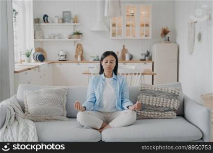 Peaceful mind, zen and concentration. Young hispanic woman is practicing yoga at home. Girl sitting on couch in lotus pose and meditating with her eyes closed. Emotion balance and stress relief.. Peaceful mind, zen and concentration. Young hispanic woman is practicing yoga at home.