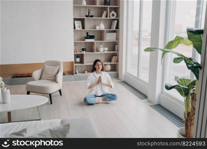 Peaceful woman practicing yoga sitting on floor in modern living room at home, keeping namaste hands gesture. Calm female breathing fresh air indoors, meditating. Mindfulness, stress relief concept.. Female meditates practicing yoga sitting on floor, breathing fresh air in modern living room at home