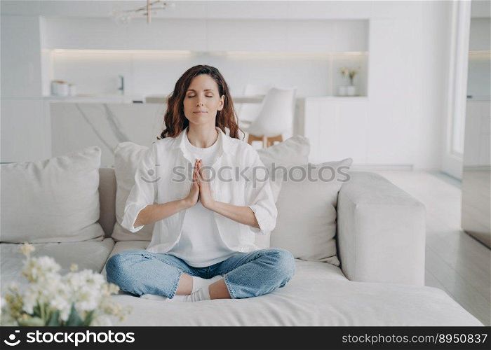 Peaceful young latin woman is practicing yoga and meditation on couch at home. Attractive girl is doing posture exercise in lotus asana. Morning gymnastics in living room. Balance, zen and harmony.. Peaceful young woman is practicing yoga and meditation on couch at home. Balance, zen and harmony.