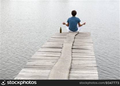 Peaceful young man doing yoga and meditation with bott≤of beer onπer at lake