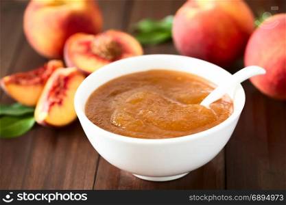 Peach jam or jelly in bowl with fresh ripe peach fruits in the back, photographed on dark wood with natural light (Selective Focus, Focus one third into the jam). Peach Jam or Jelly