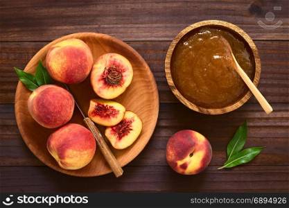 Peach jam or jelly in wooden bowl with fresh ripe peach fruits on wooden plate on the side, photographed overhead on dark wood with natural light. Peach Jam or Jelly