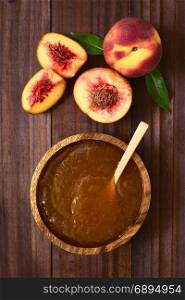 Peach jam or jelly in wooden bowl with fresh ripe peach fruits on the side, photographed overhead on dark wood with natural light. Peach Jam or Jelly