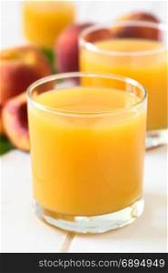 Peach juice or nectar in glasses with fresh ripe peach fruits in the back, photographed on white wood with natural light (Selective Focus, Focus on the front rim of the glass). Peach Juice or Nectar