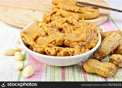Peanut butter in the bowl, peanuts in the shell and cleaned, bread, knife on the background of a linen tablecloth
