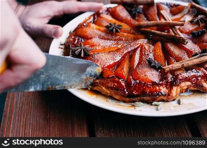 Pear pie with caramel, spices, juicy delicious tart beautifully decorated with cinnamon sticks, cardamon, star anise. Hand cutting slice with knife. Image for menu or confectionery catalog