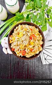 Pearl barley porridge with minced meat, tomato, yellow bell pepper, garlic and onions in a clay bowl, a towel and parsley on dark wooden board background from above