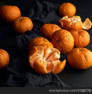 peeled ripe orange mandarin and a bunch of unpeeled round whole fruits on a black napkin, low key