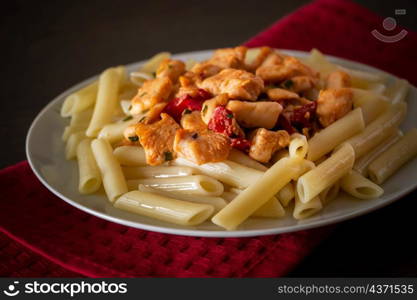 Penne pasta with chicken and vegetables in tomato sauce. Delicious penne pasta with juicy fried chicken on a plate on a red napkin, dark background. Penne pasta with chicken and vegetables in tomato sauce