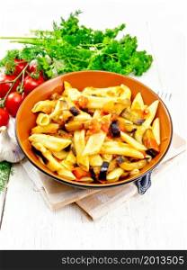 Penne pasta with eggplant and tomatoes in a bowl on napkin, fork, garlic and parsley on light wooden board background