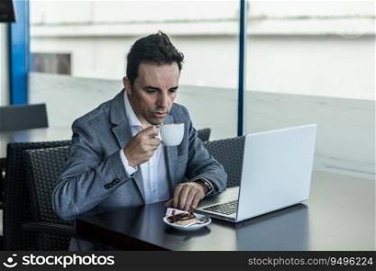 Pensive adult man in stylish formal wear drinking coffee while sitting at table with laptop in modern cafe during break. Businessman with laptop drinking coffee in cafe