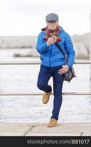 Pensive adult man wearing blue jacket and using smartphone while spending time in city park.