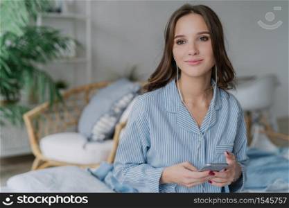 Pensive attractive woman with dark hair, hoolds modern cell phone in hands, enjoys favourite songs, uses smart phone, poses against domestic interior. Female watches online course in popular website