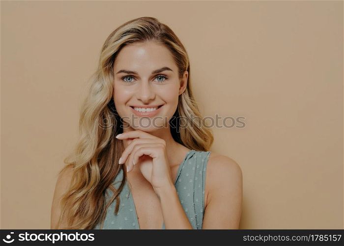 Pensive attractive young blonde female wearing blue dress holding hand under chin, smiling and looking at camera with happy face expression while standing isolated next to orange background. Attractive young blonde woman holding hand under chin