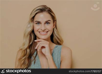 Pensive attractive young blonde female wearing blue dress holding hand under chin, smiling and looking at camera with happy face expression while standing isolated next to orange background. Attractive young blonde woman holding hand under chin