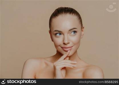 Pensive attractive young European woman with white healthy skin, looks aside, has naked body, undergoes beauty treatments, poses against beige background, has combed dark hair, bare shoulders