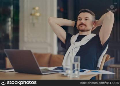 Pensive bearded man ponders on task keeps hands behind head sits at desktop with modern gadgets thinks about distance job concentrated into distance wears t shirt with sweater tied over shoulders