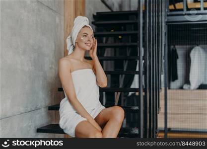 Pensive beautiful young woman applies body cream, enjoys softness of skin, sits on stairs at home, wrapped in white bath towel, looks thoughtfully into distance. Women and natural beauty concept