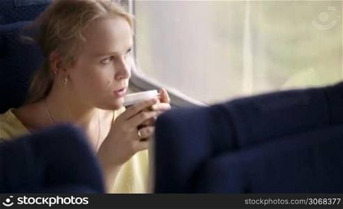 Pensive blond young married woman drinking coffee and looking out the window while traveling by train