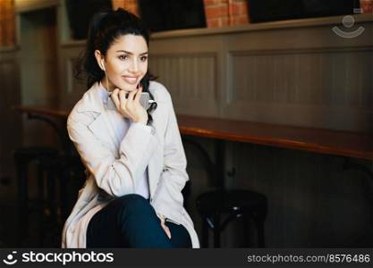 Pensive brunette female with appealing appearance looking aside while sitting in cafe enjoying favourite tracks on headphones. Relaxed woman in fashionable clothes using modern gadget indoors
