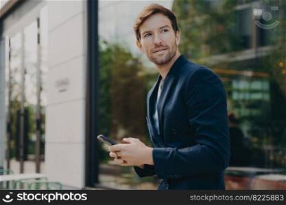 Pensive businessman holds cellular sends text messages to partner or colleague concentrated away poses near office building outdoor wears formal clothing uses easy banking app checks daily calendar. Pensive businessman holds cellular sends text messages dressed formally