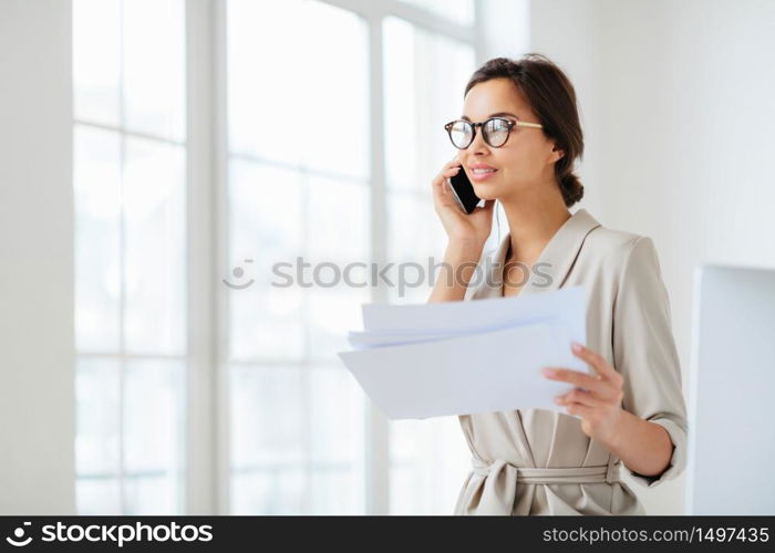 Pensive businesswoman stands sideways to camera, holds paper documents, has phone talk, prepares financial report, poses in coworking space, wears optical glasses and formal suit. Paperwork.
