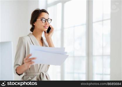 Pensive businesswoman stands sideways to camera, holds paper documents, has phone talk, prepares financial report, poses in coworking space, wears optical glasses and formal suit. Paperwork.