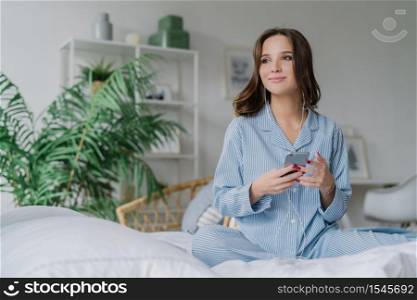 Pensive charming European woman uses app for listening music, enjoys nice sound in earphones, connected to high speed internet, spends weekend at home. Modern technologies and lifestyle concept