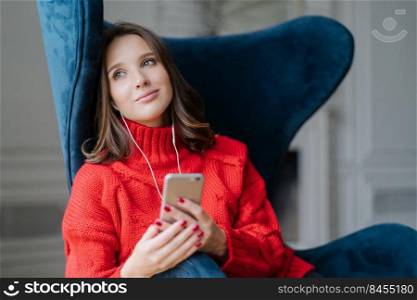 Pensive cute female feels relaxed, listens pleasant music in playlist, holds cellular, looks with dreamy expression aside, poses in comfortable armchair. People, modern technologies and coziness